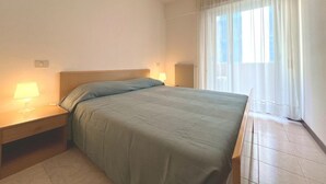 A hotel room with a bed, bedside tables, a lamp, a radiator, and a view of the outdoors through a window. - Perfect Holiday Apartment for Five Guests in Grado (Grado)
