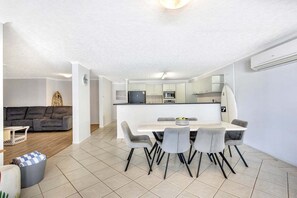 A modern kitchen with white cabinetry, a central island, and a dining area with grey chairs. - The Beach Pad - Mudjimba (Sunshine Coast)