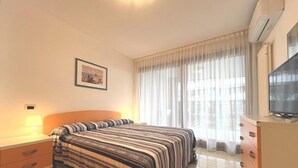 Camera d'albergo con un letto, un comodino con una lampada, una cassettiera, una televisione e una finestra con tende leggere.