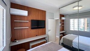 A modern bedroom with a bed, a TV mounted on the wall, a desk with a chair, and a window with a view of buildings. - New apartment in the heart of Balneário Camboriú. Comfort and sophistication in the best location. (Balneário Camboriú)