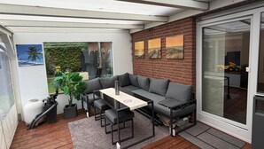 Une terrasse couverte avec un canapé, une table et des chaises, un grand bac à plantes, et une vue sur un mur en briques et une porte vitrée.