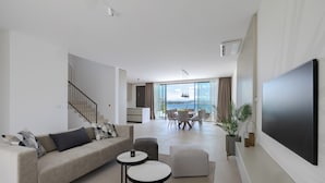 Una sala de estar moderna con televisor grande, un sofá, zona de comedor y vista al mar.