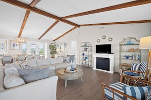 A spacious living room with a high ceiling, wooden beams, a fireplace, a television, and a dining area with wicker furniture. - Oceanfront, Direct Beach Access, Panoramic Views - 510 Tarpon Blvd (Fripp Island)