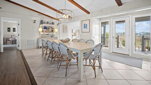 Un vaste espace dédié à la salle à manger, avec une grande table et des chaises en osier, un lustre moderne et une vue sur l’extérieur grâce à de grandes fenêtres.