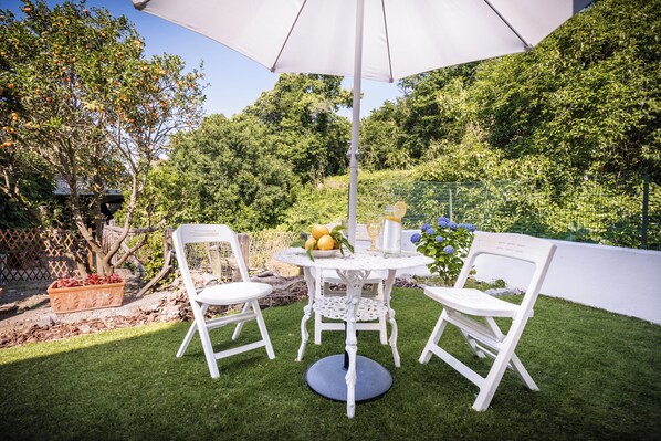 An outdoor patio with a round table, two white chairs, a white umbrella, and a potted plant with yellow lemons.
