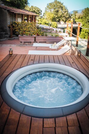 Un bain à remous sur une terrasse en bois, avec vue sur un jardin et un bâtiment en arrière-plan.