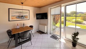 A dining area with a wooden table and chairs, a painting on the wall, a TV mounted above the table, and a view of a garden through large windows.