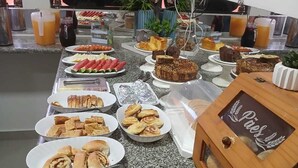 A buffet with various food items, a television displaying a landscape, and a wooden box labeled 'Paes.' - Hotéis Tropical Aquidauana (Aquidauana)