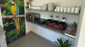 A kitchen with a microwave, toaster, and slow cooker on the counter, a shelf with plates and cups, and a parrot painting on the wall. - Hotéis Tropical Aquidauana (Aquidauana)