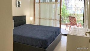 Ein Schlafzimmer mit einem Bett, einem Nachttisch, einer Lampe und einer Schiebetür zu einer Terrasse mit Stühlen.