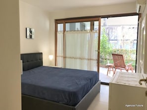 Ein Schlafzimmer mit einem Bett, einem Nachttisch, einer Lampe und einer Schiebetür zu einer Terrasse mit Stühlen.