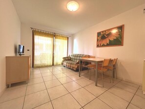 A small, well-lit room with a sofa, a dining table with chairs, a TV on a stand, and a large flower painting on the wall. - Comfortable Vacation Retreat for Four Guests (Grado)