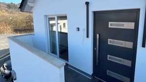 Et moderne hus i to etager med hvide facader, en mørk hoveddør med tre håndtag, en skydedør i glas og en velkomstmåtte.