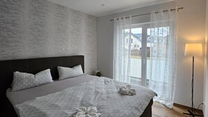 Et moderne soveværelse med en stor seng, en sengegavl, et natbord, en lampe og udsigt gennem et vindue med lette gardiner.