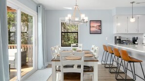 A modern kitchen with white cabinets, a dining area with a wooden table and white chairs, and a bar area with stools. - Ripple| Golf Cart · Private Beach · Outdoor Living (Emerald Isle)