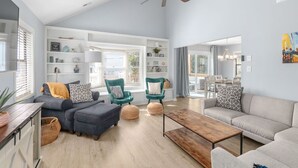 A spacious living room with a high ceiling, a ceiling fan, and a large window providing natural light. There are two sofas, a coffee table, and a bookshelf filled with books and decorative items. - Ripple| Golf Cart · Private Beach · Outdoor Living (Emerald Isle)