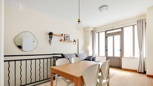 Ένας χώρος τραπεζαρίας με ένα ξύλινο τραπέζι και λευκές καρέκλες, έναν στρογγυλό καθρέφτη και ένα φωτιστικό που κρέμεται από την οροφή.