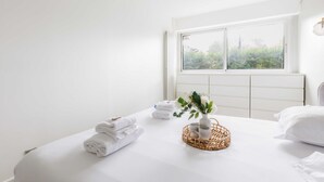 Habitación de hotel con un ventanal grande, una cama bien hecha con ropa blanca y una pequeña cesta de mimbre con un florero de flores sobre la cama.