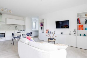 Een moderne woonkamer met een witte bank, een eethoek met tafel en stoelen, een keuken met witte kastjes en een tv aan de muur.