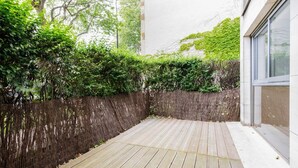 Una terraza de madera con un muro blanco y una ventana a la derecha.