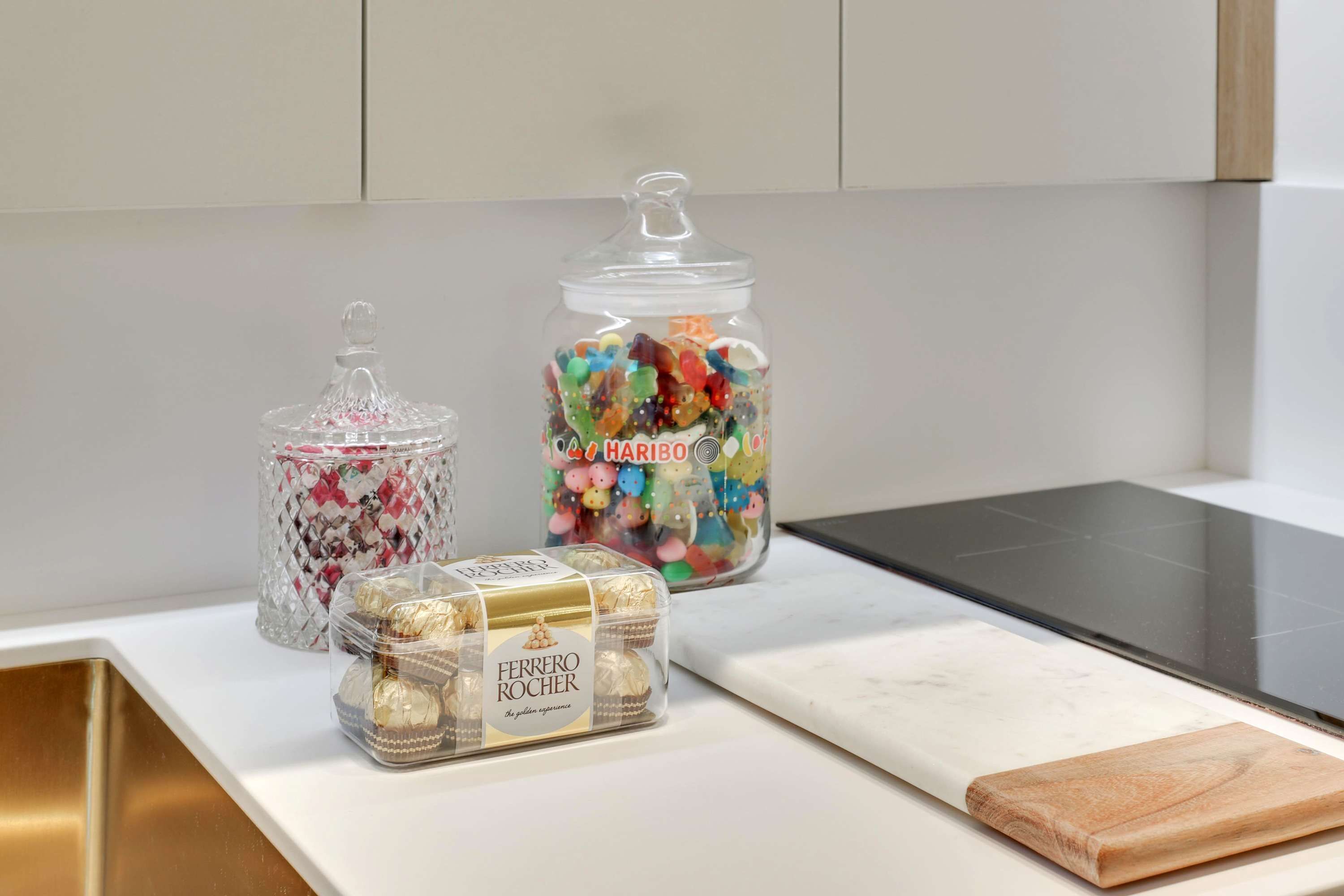 A kitchen counter with a marble top, a wooden cutting board, and three glass jars containing Ferrero Rocher chocolates and colorful candies.