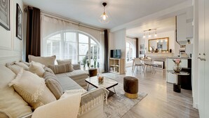 Amplia sala de estar con sofá, mesa de centro y reposapiés, zona de comedor con mesa y sillas, y cocina con espejo y mostrador.