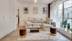 Una sala de estar con sofá, mesa de centro y dos mesitas auxiliares. Se observan cuadros en la pared y un ventanal con plantas en el alféizar.