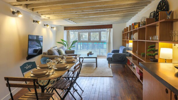 Un coin repas avec des meubles en bois, un téléviseur fixé au mur et une vue sur l’extérieur par une fenêtre.