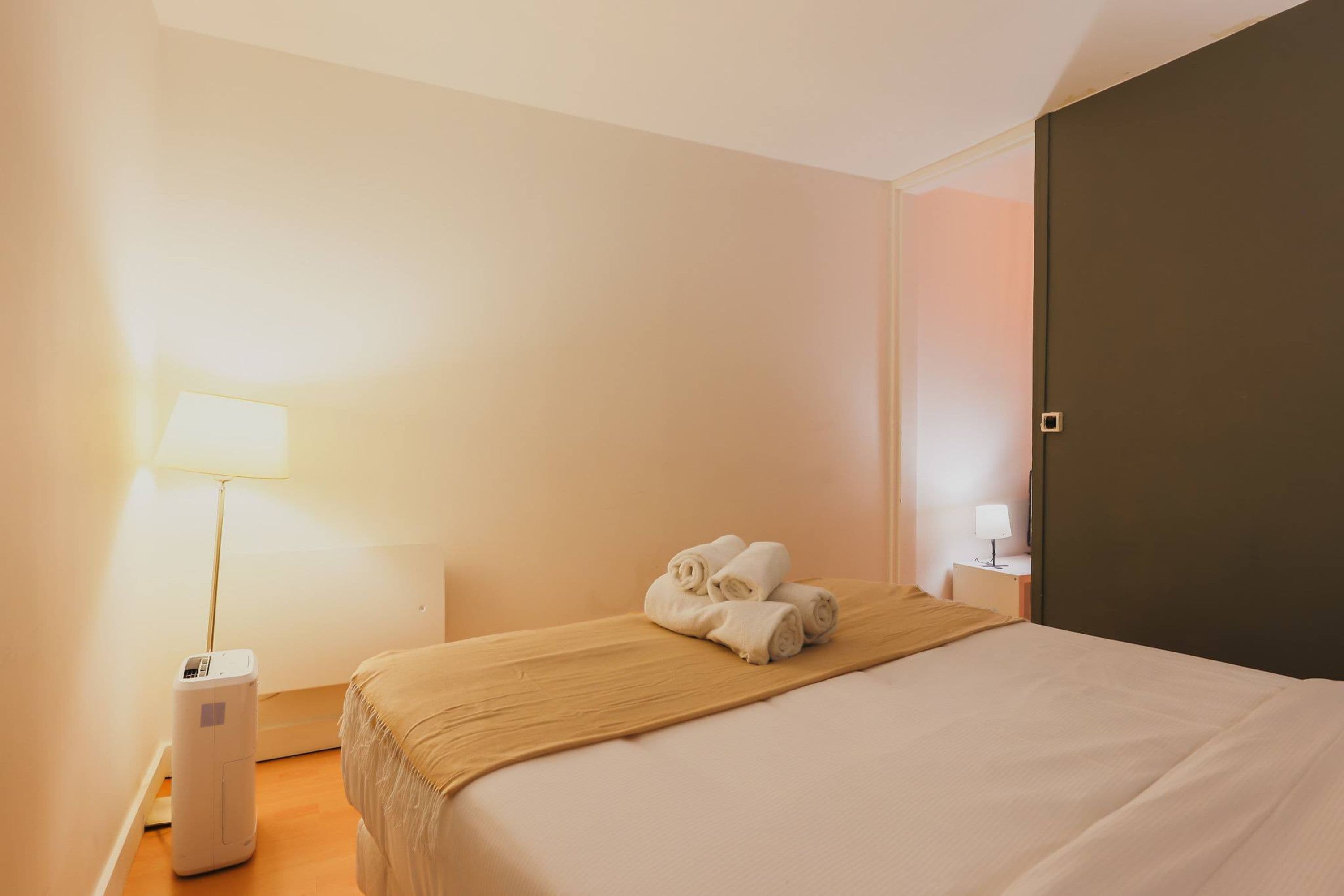 A hotel room with a bed, a bedside table with a lamp, a heater, and rolled towels on the bed.
