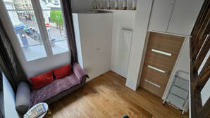 Een knus kamertje met raam, een bank met kussens, een klein tafeltje met een tijdschrift, en een houten vloer.