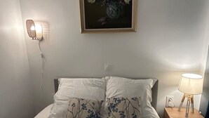 A neatly made bed with white linens and floral-patterned pillows, a framed floral painting above it, a wall-mounted lamp, and a bedside table with a lamp. - Flat Near Notre Dame 4 pax AC (Paris)