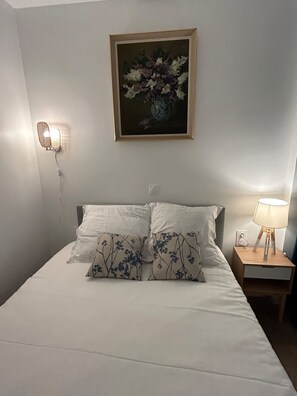 Ein ordentlich bezogenes Bett mit weißen Leinentüchern und Kissen mit Blumenmuster, ein gerahmtes Blumenbild darüber, eine Wandlampe und ein Nachttisch mit Lampe.