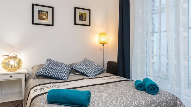 A neatly arranged hotel room with a bed, two framed pictures on the wall, a bedside lamp, and a window with sheer curtains. - Place des Vosges Havre de Paix Paris 8 Pers AC (Paris)