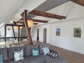Un loft amplio con techo alto, vigas de madera a la vista, un sofá gris con cojines estampados y una escalera que conduce a un nivel superior.