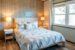 A bedroom with a wooden wall, a bed with a patterned bedspread, two bedside tables with lamps, and a window with blinds. - Private Beach Front: Rustic Dune Retreat (St. Augustine)