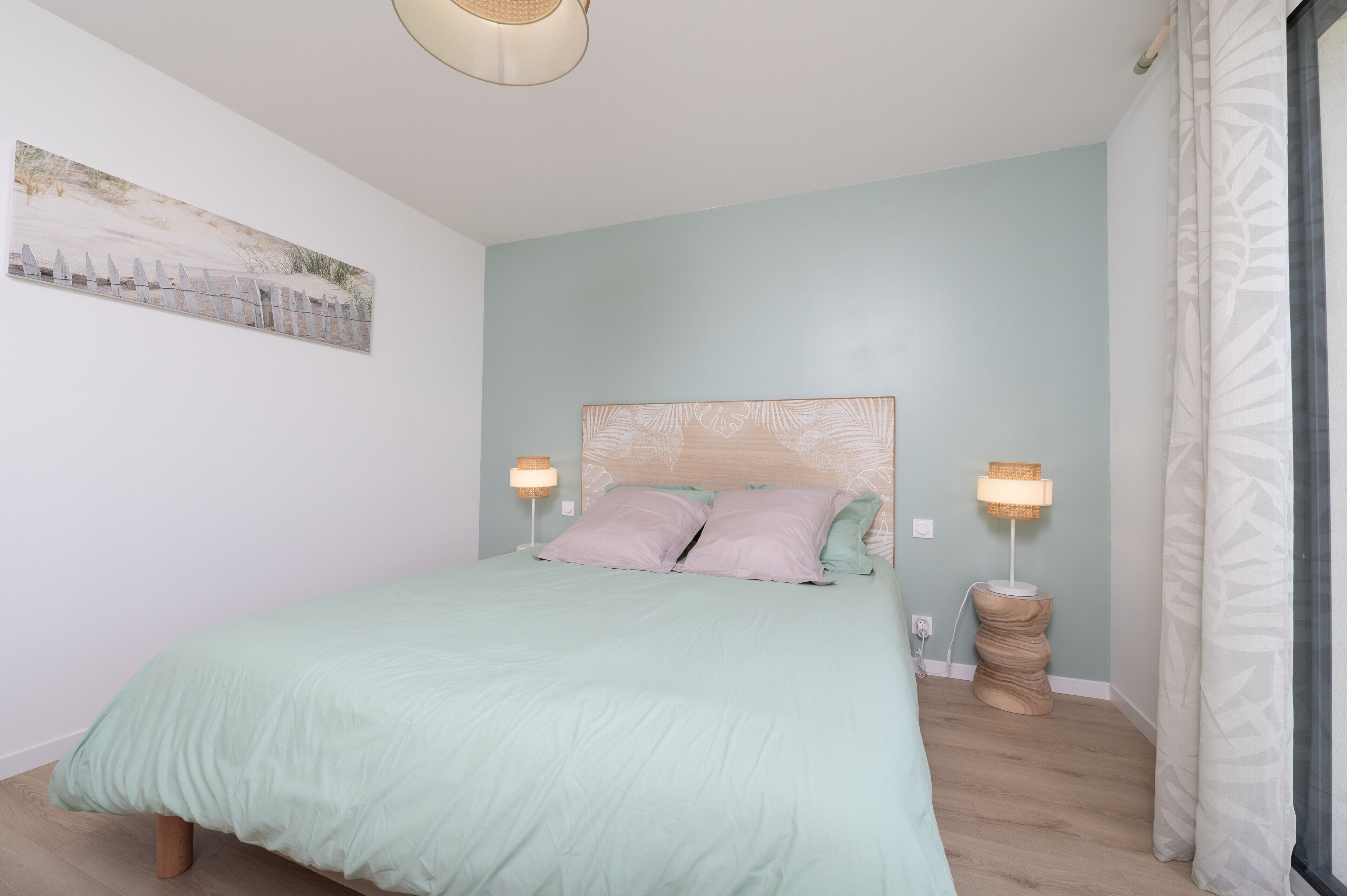 A bedroom with a bed, two bedside lamps, a wall art piece, and a window with curtains.