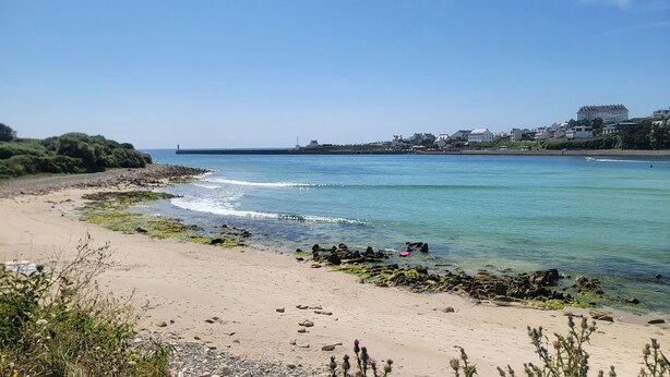 Une plage côtière aux eaux cristallines, avec un rivage rocheux et des bâtiments visibles à l’arrière-plan.