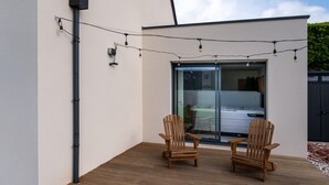 Une terrasse en bois avec deux chaises et des guirlandes lumineuses, une porte-fenêtre coulissante et un véhicule à l’intérieur.