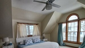 A bedroom with a bed, a ceiling fan, a nightstand, a window with curtains, and a view of the outside.