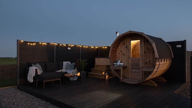 Un sauna en forme de tonneau en bois, avec un banc et une petite table, entouré de guirlandes lumineuses sur une terrasse en bois.