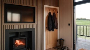 Une pièce avec un mur lambrissé, une cheminée où l’on voit les flammes, une porte sur laquelle sont accrochés des manteaux, et une vue sur un paysage verdoyant par une porte vitrée.