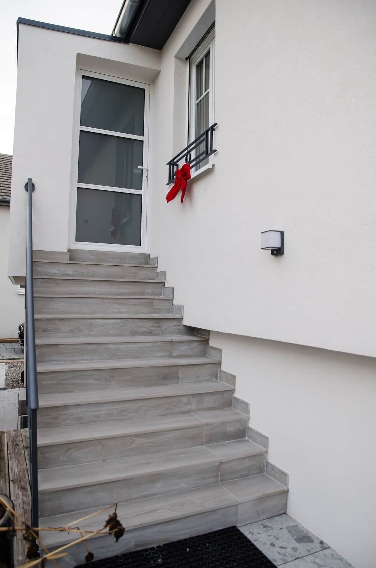 Eine Treppe führt zu einer weißen Tür mit einem roten Band, einem Fenster mit schwarzem Rahmen und einer kleinen Wandleuchte.
