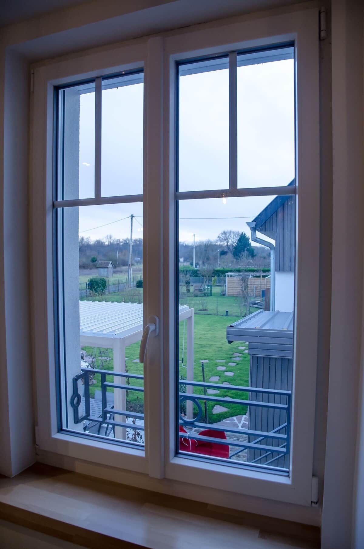 Une fenêtre donnant sur un jardin avec une pelouse, une terrasse en bois et un abri de jardin.