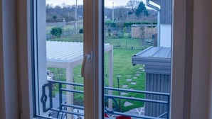 Une fenêtre donnant sur un jardin avec une pelouse, une terrasse en bois et un abri de jardin.