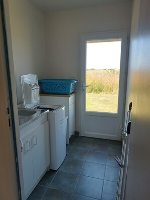 A compact kitchen with a washing machine, a small sink, and a blue laundry basket. - Ti Mammig. (Kerlouan)
