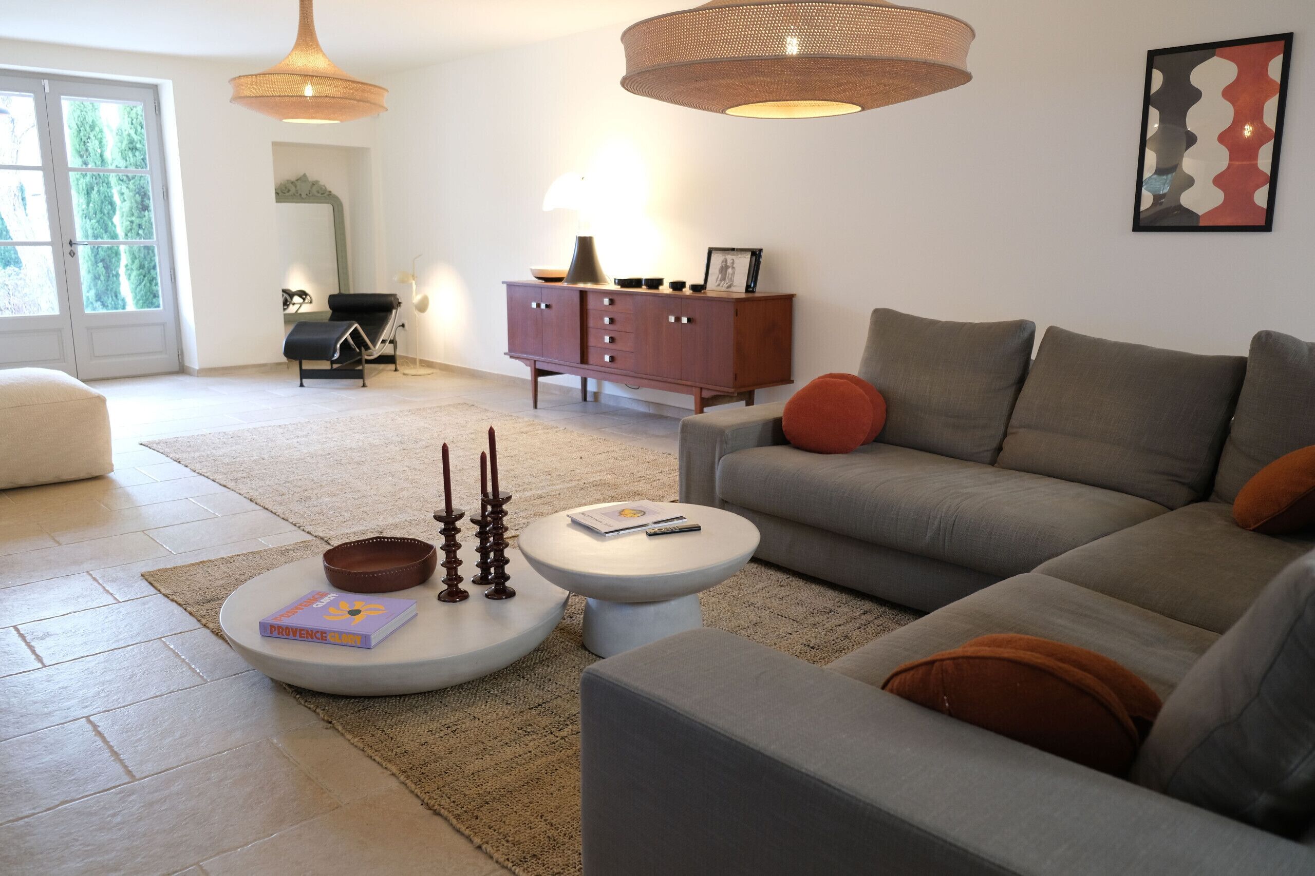A modern living room with a grey sofa, a white coffee table, and a brown leather ottoman. There is a wooden cabinet with a lamp, a framed picture, and a small table with candles.