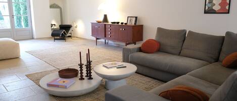 A modern living room with a grey sofa, a white coffee table, and a brown leather ottoman. There is a wooden cabinet with a lamp, a framed picture, and a small table with candles.