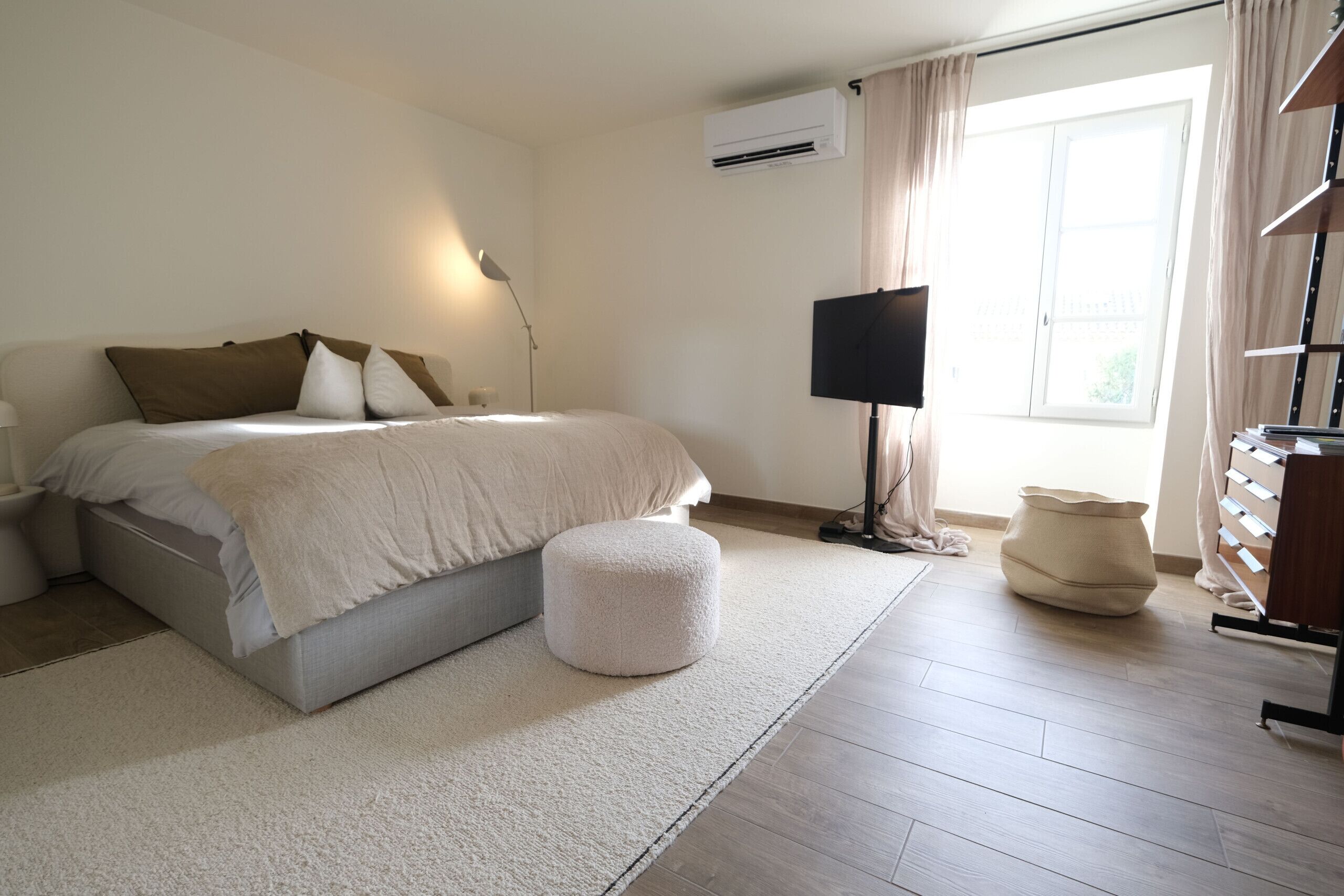 A bedroom with a bed, a bedside table, a lamp, a TV, and a window with curtains.