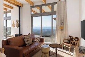 Una sala de estar moderna con un sofá de cuero marrón, una silla de madera y una mesa redonda de madera. Un ventanal ofrece vistas a las montañas.