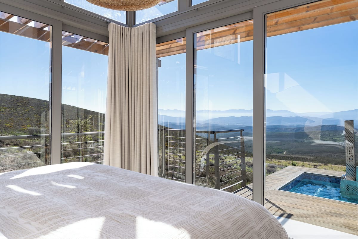 A bedroom with a large window overlooking a mountainous landscape, a wooden deck, and a hot tub.